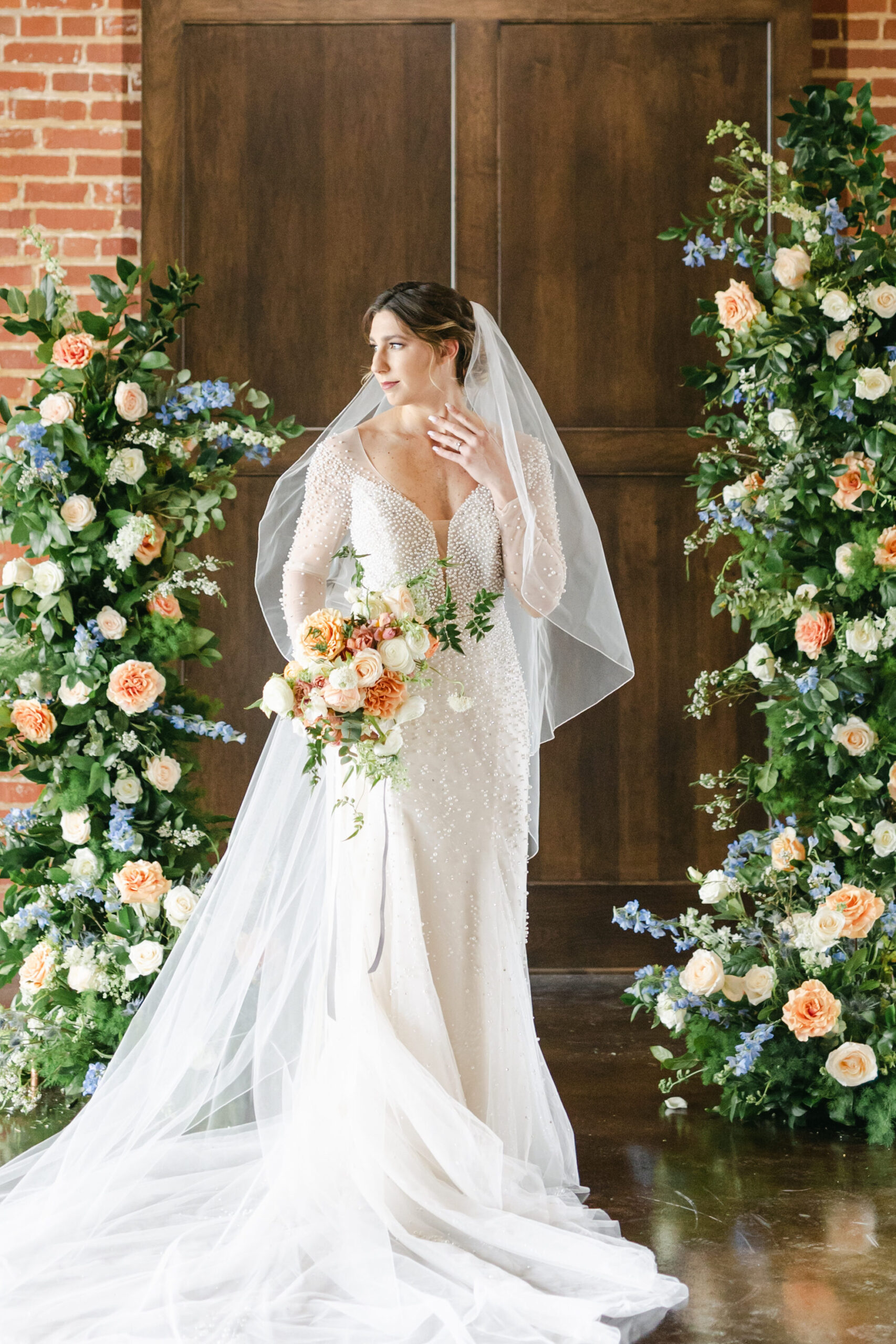Summer 2023 bride with blue and peach floral arch holding a whimsical style peach bouquet