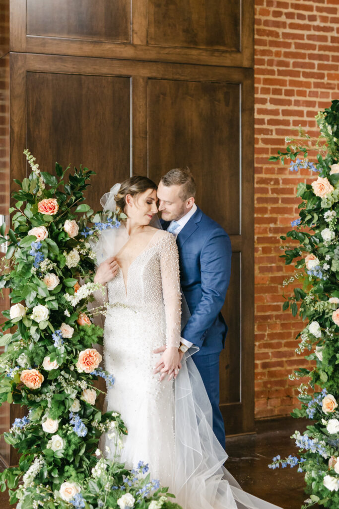 Summer 2023 bride and groom smiling in embrace with floral arch with white and blue flowers 